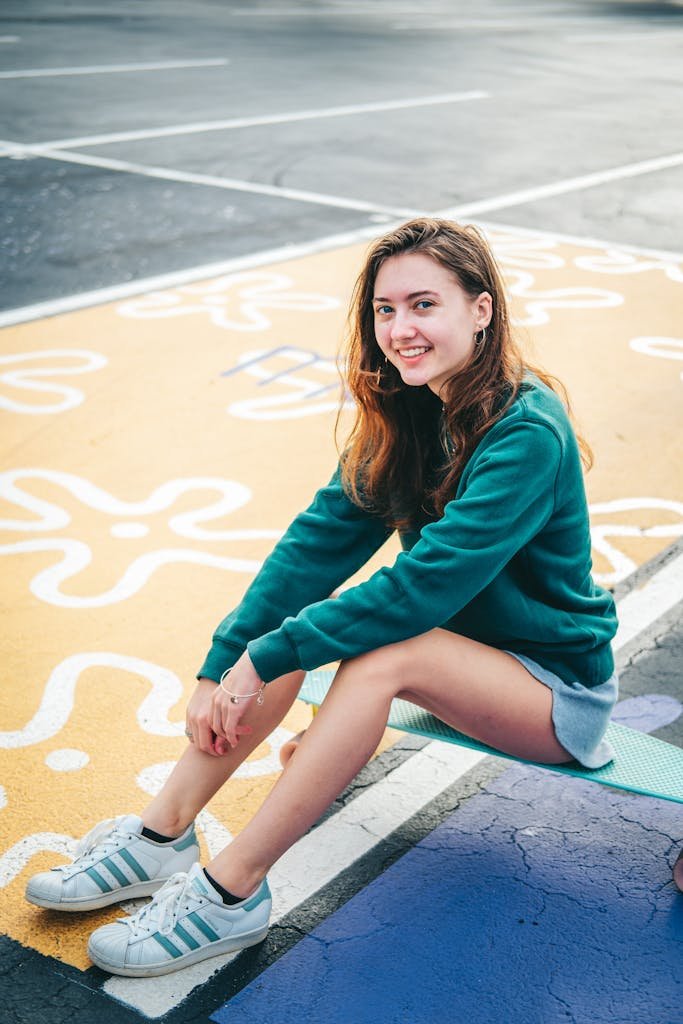 A young woman with long hair smiles while sitting on a skateboard in a vibrant outdoor setting.