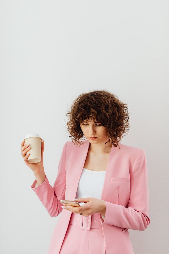 Confident woman in a pink blazer holding coffee and texting indoors.