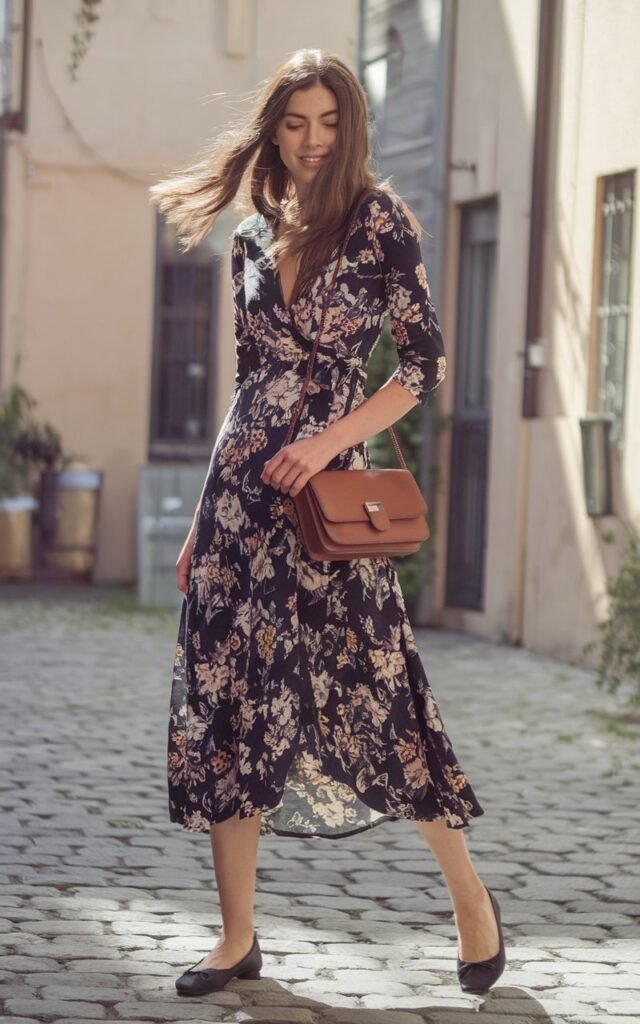 A model in a floral midi wrap dress styled with classic black ballet flats and a crossbody bag. Shot in a cobblestone alley with soft natural daylight. Her hair flows naturally in the breeze, expression warm and effortless.