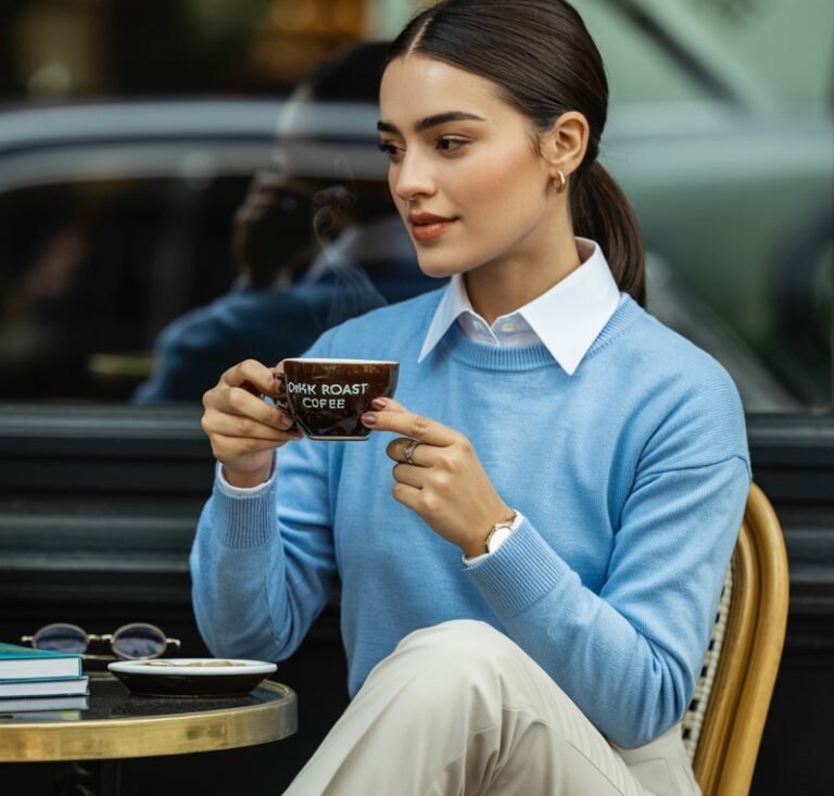 A model in a pale blue crewneck sweater layered over a crisp white button-down, paired with slim beige trousers and polished brown loafers. Shot in a quiet café with window light creating soft shadows across her face. She sits casually on a wooden chair, hair in a sleek ponytail, looking thoughtful while stirring coffee.