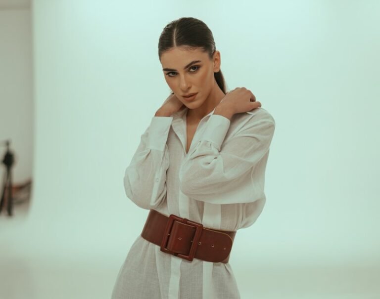 A model poses against a minimal white studio wall under soft indoor light. She wears a crisp oversized shirt dress cinched with a wide leather belt, paired with knee-high boots. Hair is sleek in a high ponytail, expression strong but playful.