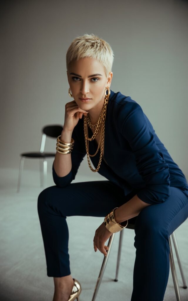 A short-haired blonde with sharp features poses in a modern indoor studio with warm spotlighting. She wears a fitted navy jumpsuit styled with chunky layered gold chains, bangles, and metallic heels. Sitting on a stool, leaning slightly forward with an elegant, powerful gaze.