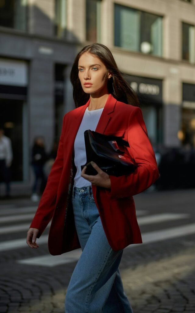 A tall brunette model with piercing blue eyes walking through Milan’s fashion district, wearing a vibrant red tailored blazer, white tee, and high-waisted denim, carrying a luxury leather tote, dramatic side lighting, modern power-woman vibe.