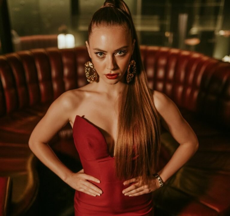 A white-skinned brunette with a sleek ponytail poses in a dimly lit lounge. She wears a curve-hugging deep red bodycon dress with oversized gold statement earrings and strappy stilettos. Standing tall with hands on hips, giving a confident, unstoppable expression.