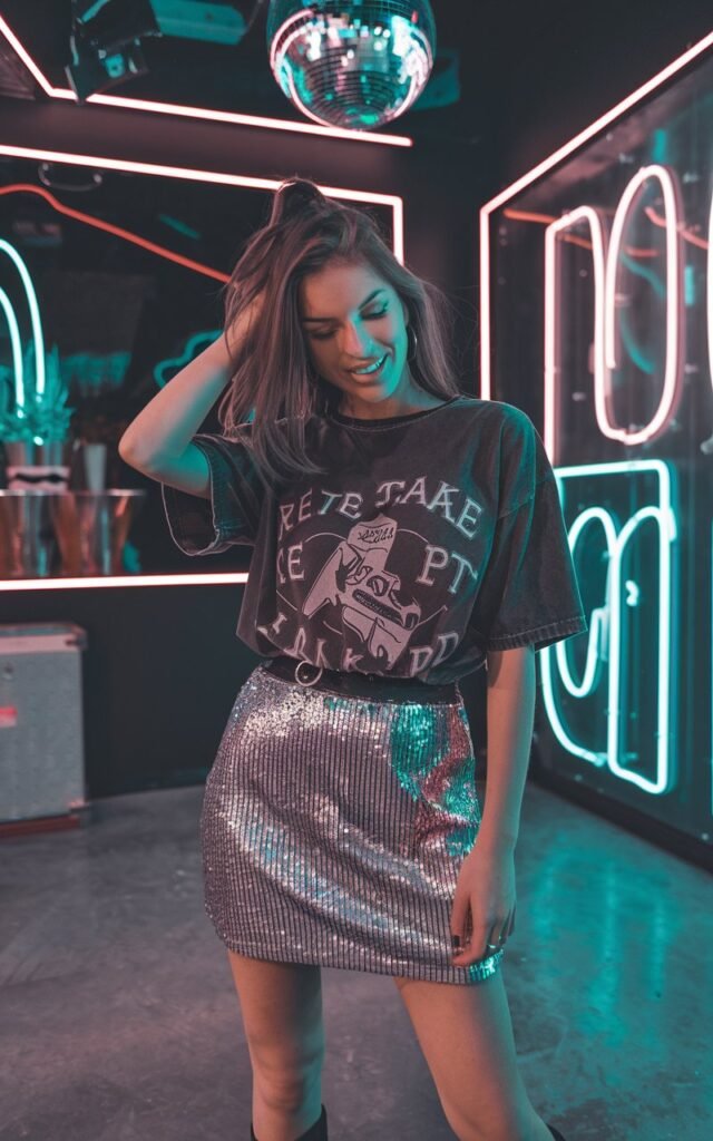 An indoor nightclub-inspired studio setup with neon lighting. The model wears a silver sequin mini skirt with a black graphic tee tucked in, styled with ankle boots. Playful pose, one hand through her hair, smirking at the camera.