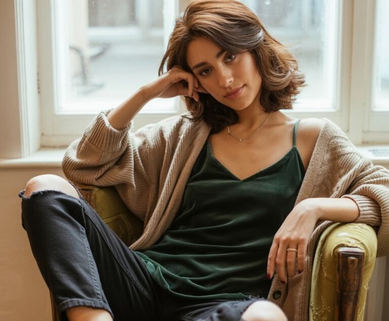 Industrial warehouse setting with moody daylight streaming in. Brunette model with bangs wears a black mesh long-sleeve under a baggy oversized graphic tee, paired with distressed shorts and chunky boots. She leans against a concrete pillar, arms crossed, expression rebellious yet playful.