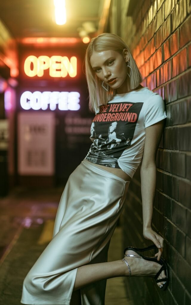 Editorial-style image of a tall model with straight blonde hair, wearing a silver satin midi skirt, tucked vintage band graphic tee, and strappy black heels. Setting is a brick-wall alleyway with neon signs just turning on at dusk (golden hour glow). She’s leaning casually against the wall, knotting her tee at the waist, with a confident smirk and bold earrings catching the light.