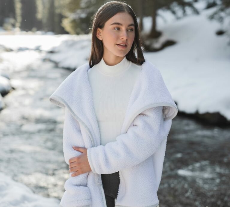 A young model stands by a snow-covered stream with soft daylight. She wears a white thermal bodysuit under a fluffy sherpa coat, paired with grey leggings and snow boots. She stands with one arm wrapped around herself, eyes sparkling with warmth. The winter landscape and soft lighting bring out the cozy textures and natural beauty of the scene.