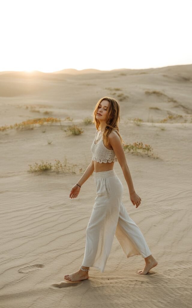 Boho-inspired shoot in a desert location during golden hour. Model wears a white crochet crop top with high-rise wide-leg flowy trousers and leather sandals. Hair loose and windblown. She walks barefoot in the sand, dreamy and free-spirited.