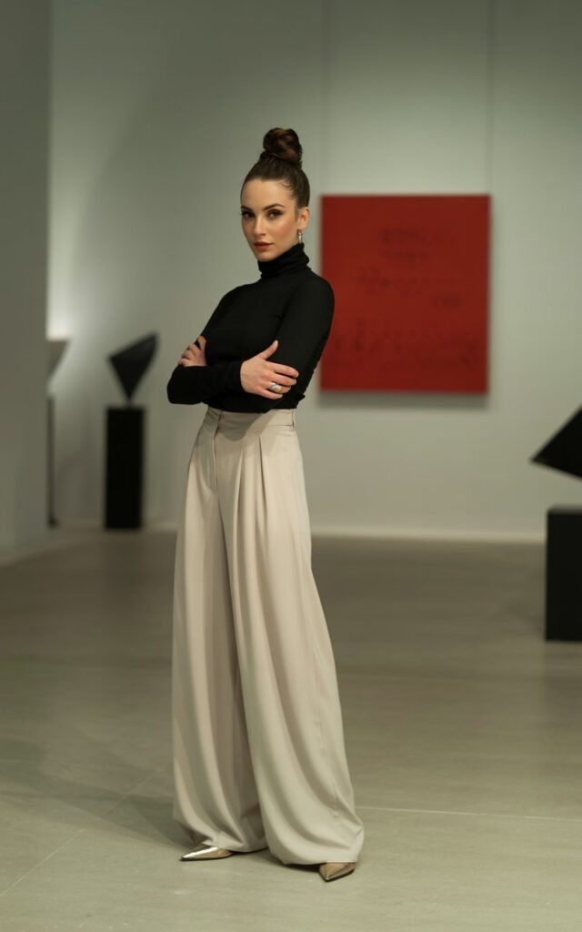 Elegant brunette with sleek bun, wearing a black fitted turtleneck, cream palazzo pants, and pointed flats. Shot in a minimalist art gallery, soft indoor light. She’s standing with arms crossed, chic and poised.