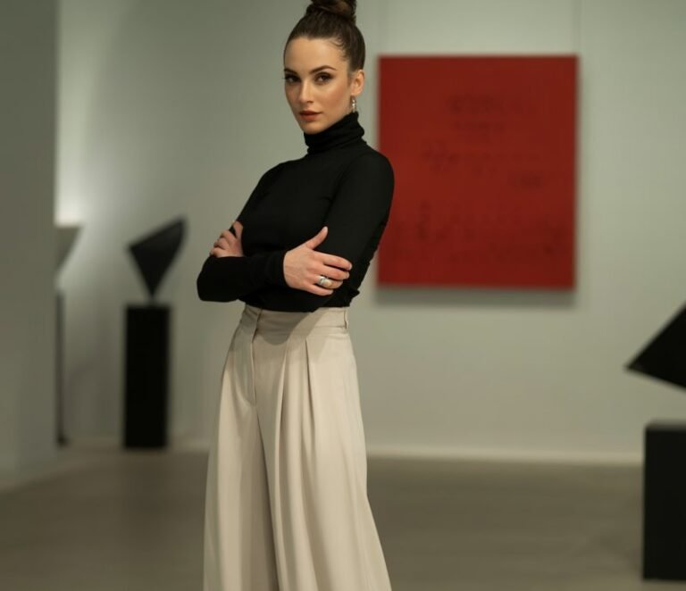 Elegant brunette with sleek bun, wearing a black fitted turtleneck, cream palazzo pants, and pointed flats. Shot in a minimalist art gallery, soft indoor light. She’s standing with arms crossed, chic and poised.