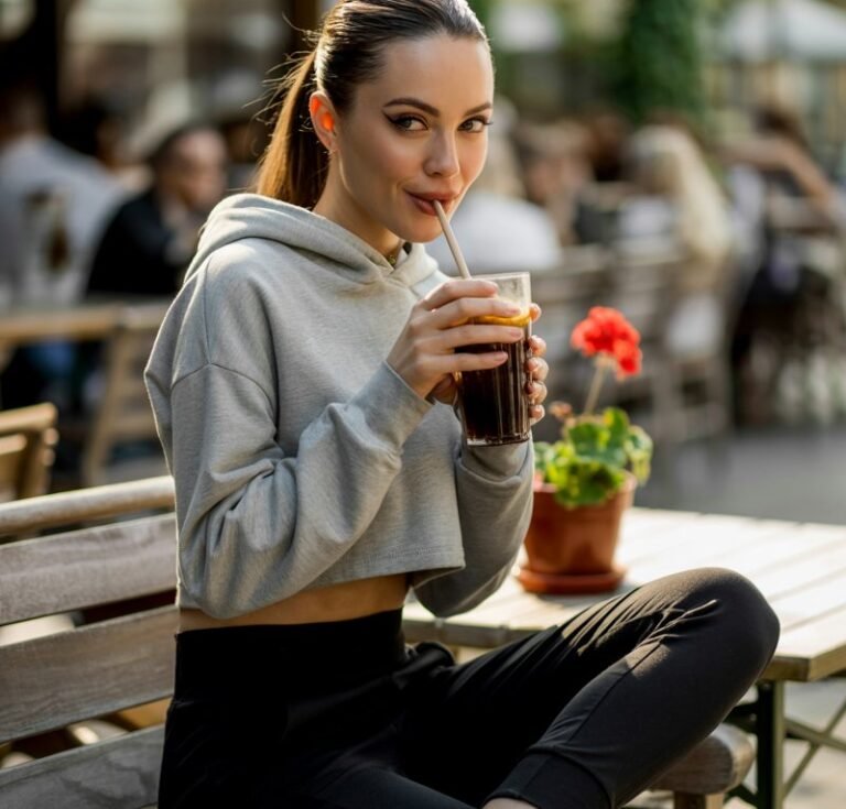 Full-body image of a white female model in a cropped gray hoodie and fitted high-waisted black joggers with white sneakers. She’s sitting on a café bench outdoors, sipping iced coffee, golden-hour sunlight highlighting her sharp jawline. Hair pulled into a sleek high ponytail, expression playful yet chic.