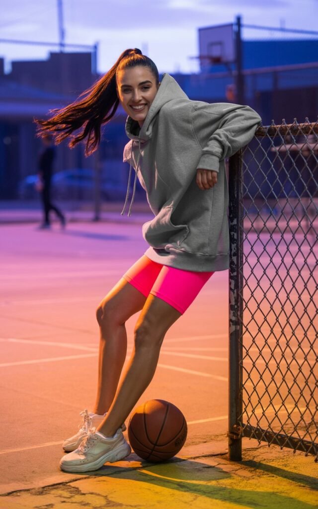 Full-body sporty photo of a brunette model with long ponytail, wearing neon pink bike shorts, oversized gray hoodie, and chunky sneakers. Captured in an urban basketball court at sunset, vibrant natural light. She’s leaning against a fence, relaxed with a playful smirk.