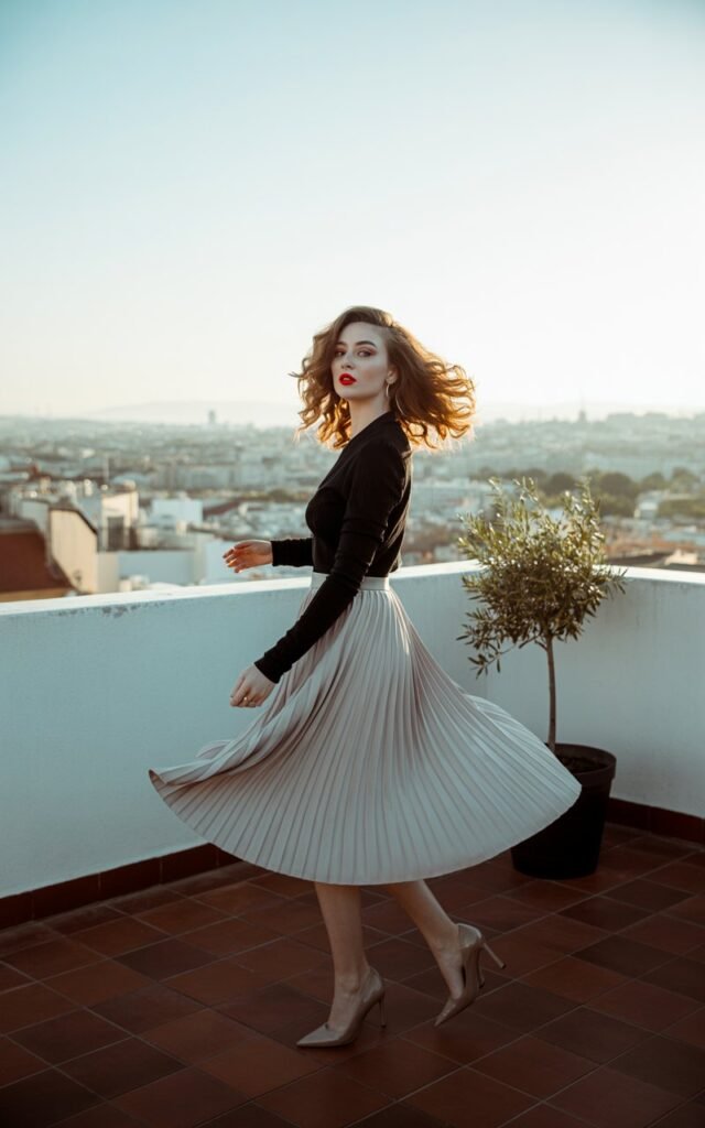 Full-body urban rooftop photo. Model wears a flowy pleated white midi skirt with a slim black long-sleeve top tucked in. Pointed heels and red lipstick finish the look. Hair styled in loose glamorous curls. Golden-hour lighting enhances elegance as she twirls lightly, skirt flowing.