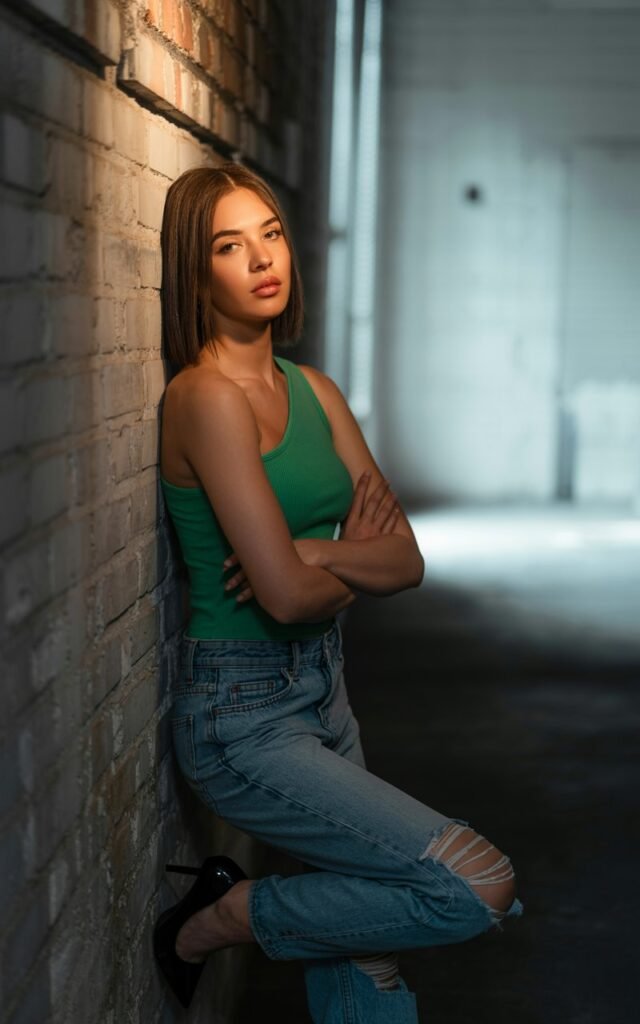 Industrial warehouse backdrop with soft window lighting. Model wears a fitted one-shoulder top in bold color, paired with relaxed ripped boyfriend jeans and heels. Hair straight and sleek. Pose leaning against a wall, arms crossed, edgy and confident.