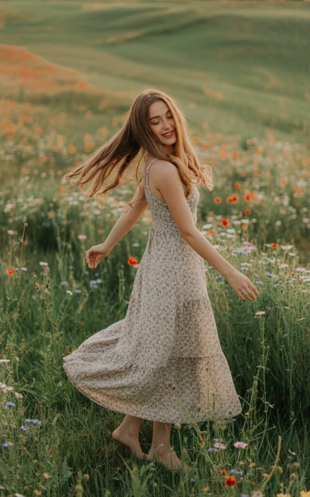 Nature setting with wildflowers during golden hour. Model wears a flowing maxi dress in muted floral prints with bare feet in the grass. Hair in soft waves. Warm light catches the fabric as she spins playfully, carefree and romantic.