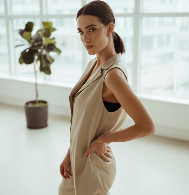 Shot in a minimalist loft space with floor-to-ceiling windows. Model wears a beige sleeveless cotton jumpsuit with black slide sandals. Hair in a sleek low ponytail. Natural window light highlighting fabric texture. She stands tall with one hand on her hip, confident and chic.