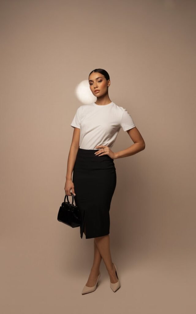 Studio full-body shot on a minimal beige backdrop. A model stands with one hand on hip, wearing a plain white tee tucked into a sleek black pencil skirt. She finishes with nude pumps and a structured handbag. Her hair is styled sleek and straight, soft glam makeup. Studio lights highlight clean contours as she poses with confident elegance.