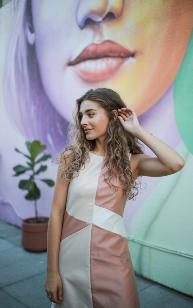 The model poses in front of a modern art mural painted with soft pastel hues. She wears a color-block shift dress in shades of cream and muted pink with geometric panels. Her hair is in loose waves, flowing naturally around her face. Soft daylight illuminates the space, creating gentle shadows. She stands with one hand brushing her hair back, smiling softly, looking comfortable and modern.