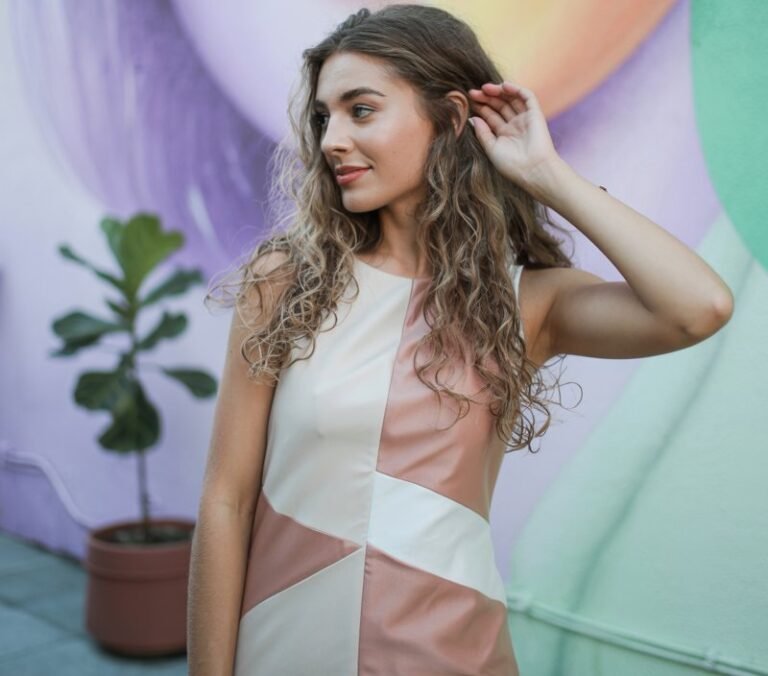 The model poses in front of a modern art mural painted with soft pastel hues. She wears a color-block shift dress in shades of cream and muted pink with geometric panels. Her hair is in loose waves, flowing naturally around her face. Soft daylight illuminates the space, creating gentle shadows. She stands with one hand brushing her hair back, smiling softly, looking comfortable and modern.
