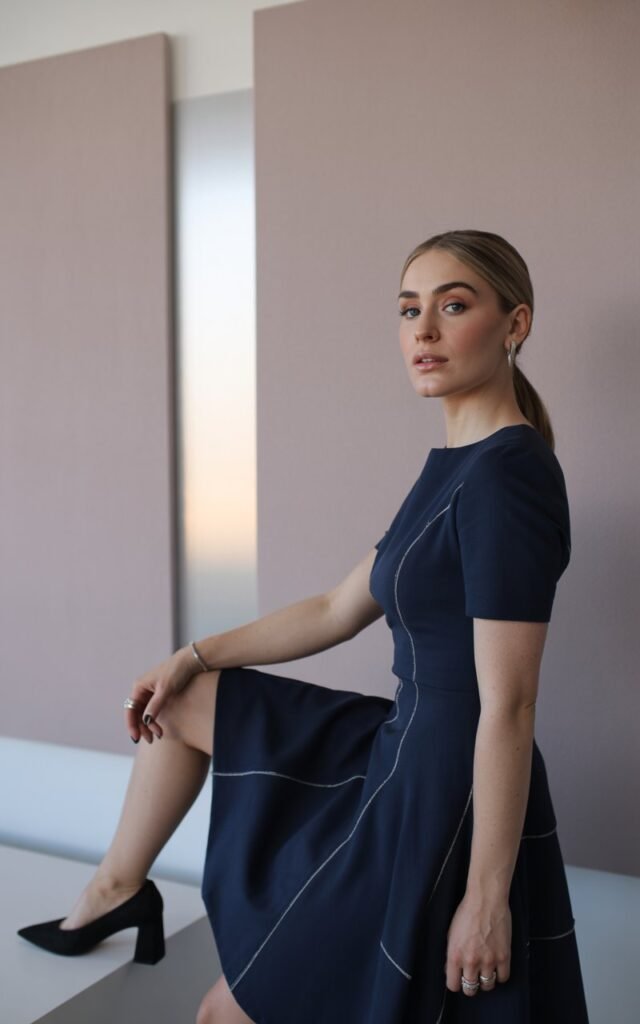 The model stands confidently in front of a modern art gallery wall painted in neutral tones. She wears a navy blue fit-and-flare dress with sharp seam detailing that contours her waist and hips. Her hair is pulled back into a sleek ponytail, and soft makeup enhances her sharp cheekbones. She pairs the dress with block heels and minimal rings. Her expression is poised, with one leg slightly forward, radiating elegance.