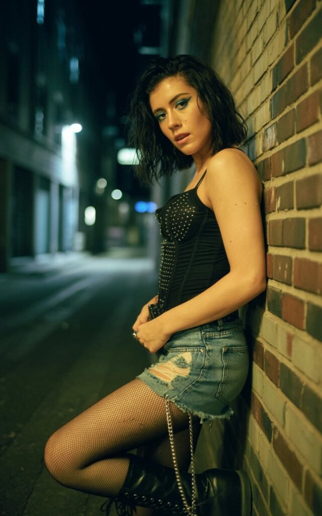 Urban alleyway setting at night with moody shadows. Model wears a black studded bustier, ripped denim shorts layered over fishnet tights, and lace-up boots. She stands leaning against a brick wall, chin slightly lifted, smoldering expression. Dark tousled hair, smoky eyeliner. A fierce and rebellious energy.