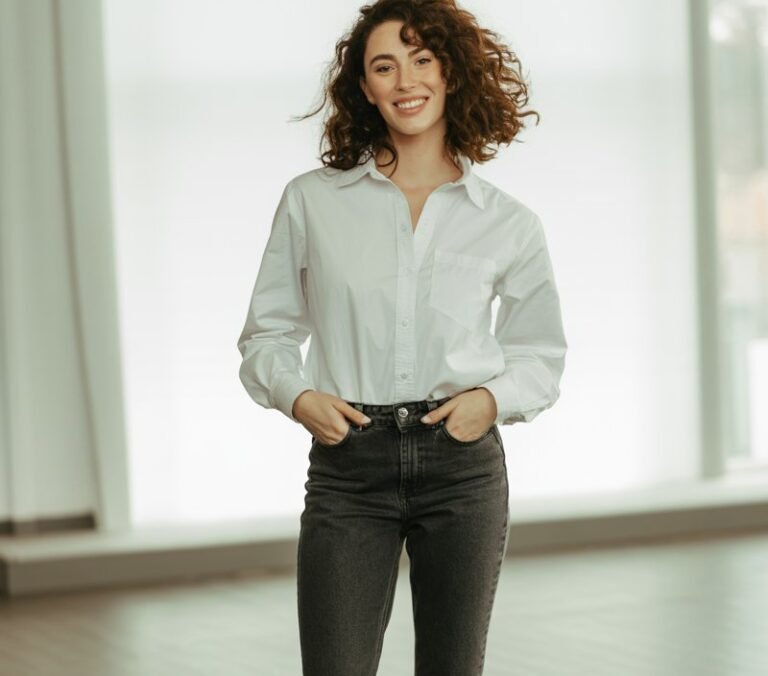 White-skinned model with natural brunette waves, wearing dark wash skinny jeans, crisp white tucked button-up, and tan loafers. Captured in a sunlit modern studio with wooden flooring. She’s standing tall, hands tucked in pockets, smiling naturally, giving timeless simplicity.