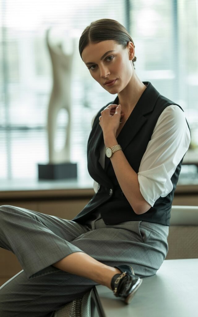 A white-skinned woman with sleek dark hair poses in a modern office setting. She wears a tailored black vest layered over a white shirt and gray trousers. Simple accessories and pointed loafers complete the look. Natural indoor light through blinds adds subtle texture and contrast.