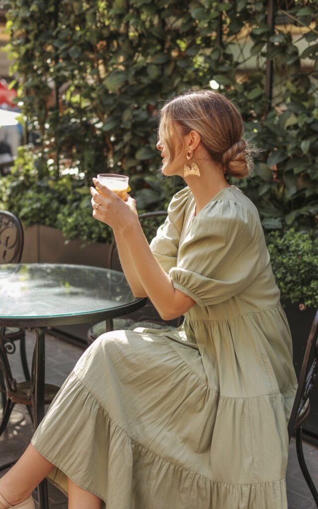 Captured in a sunlit café patio scene. The model sits elegantly with a drink, wearing a sage green tiered midi dress and chunky gold earrings. Her hair is styled in a low messy bun with soft strands framing her face. Natural daylight filters through greenery, highlighting the texture of her dress.