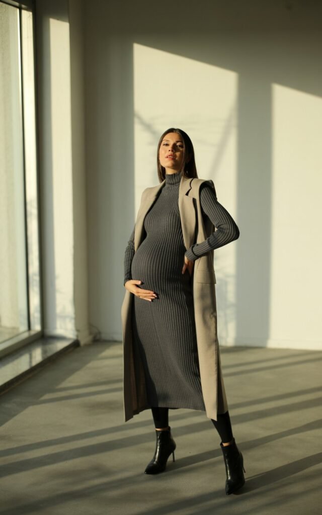 Elegant pregnant woman in a ribbed charcoal midi dress with a long beige vest. Indoor minimalist space with large windows, soft directional light. Standing tall, hair sleek and tucked behind ears, wearing heeled boots.