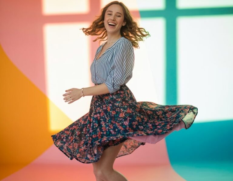Full-body image in a vibrant studio with colorful backdrops and soft natural lighting. The model wears a striped blouse with a floral skirt, colorful heels, and minimal jewelry. Her hair is loose and natural, and she twirls mid-motion, smiling with joy.