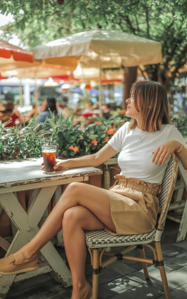 Full-body outdoor shot in a garden café. The model wears a white knit top tucked into high-waisted beige paperbag shorts and tan espadrilles. She’s seated at a wooden table with iced coffee, natural sunlight glowing on her skin. Expression relaxed and content.