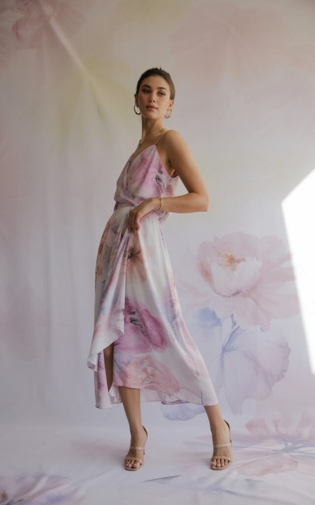Full-body view in a pastel studio backdrop. The model wears a watercolor floral wrap dress, nude sandals, and minimal jewelry. Soft natural lighting highlights the print’s delicacy. She’s posed elegantly with one hand resting on her hip.