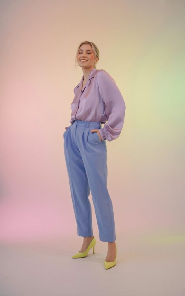 Full-body view in a pastel-toned studio with soft lighting. The model wears a lavender blouse, baby-blue trousers, and pale-yellow heels. Her makeup is dewy and minimal, and she’s smiling confidently with one hand in her pocket. The pastel background complements the outfit.