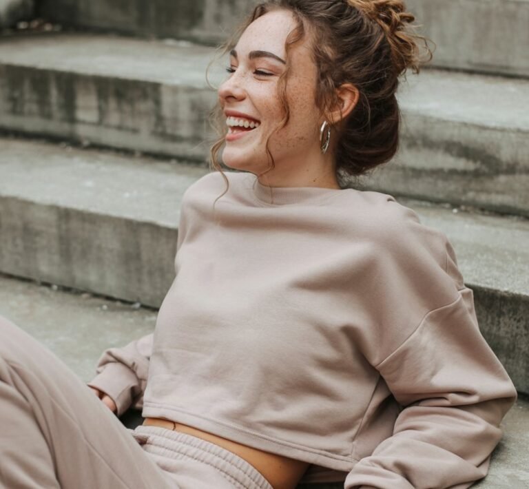Model with natural freckles and light brown hair in a messy bun lounges on concrete steps. She wears a soft beige cropped sweatshirt with matching joggers and white sneakers. Soft daylight, cozy texture emphasis. Relaxed posture with a candid laugh — effortlessly cool.