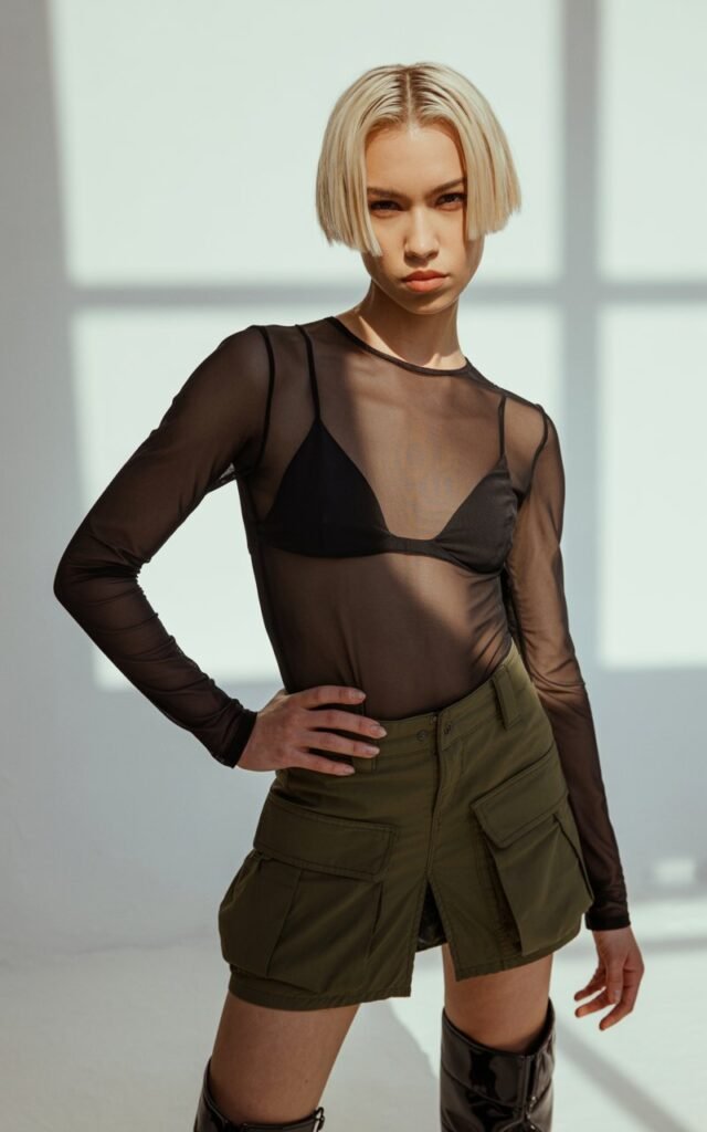 Model with platinum-blonde bob poses under soft studio lighting. She wears a sheer black long-sleeve top over a bralette, olive cargo skirt, and chunky statement boots. Textured fabrics and realistic skin detail. Fierce stance, one hand on hip — confident and bold.