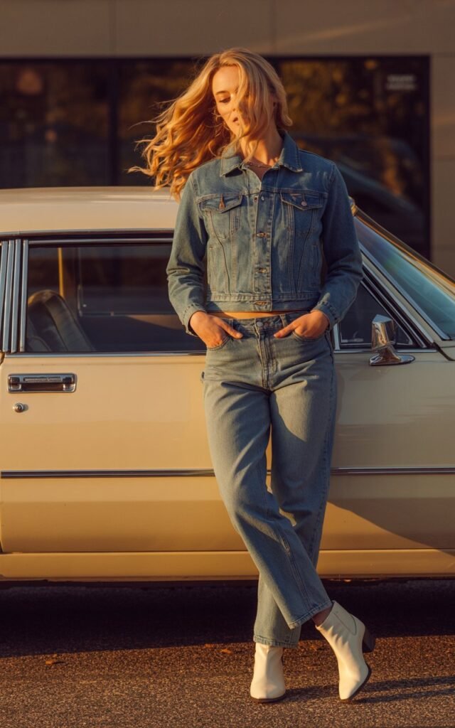 Model with wavy blonde hair stands beside a vintage car. She wears a fitted medium-wash denim jacket, relaxed jeans, and white ankle boots. Late-afternoon sunlight enhances the denim tones. Casual pose with hands in pockets, hair moving in the breeze, cinematic realism.