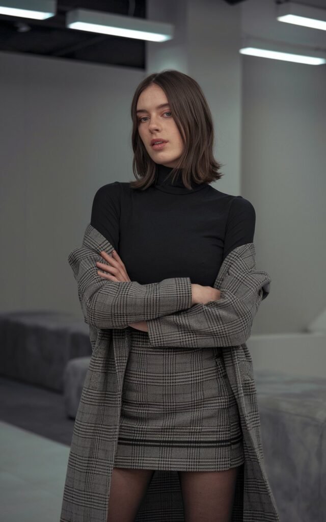 Modern indoor photo of a white-skinned woman with shoulder-length brunette hair wearing a black fitted turtleneck, plaid skirt, and sheer tights. Minimal studio setting with soft diffused lighting. She’s standing confidently with arms crossed, slight smirk on her lips.