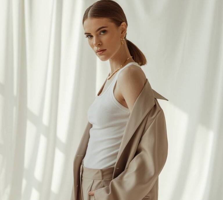 Studio editorial setup with diffused lighting. Model stands tall in a white tank layered under a beige oversized blazer with matching trousers. Minimal accessories, slick low ponytail. Pose slight lean forward, confident gaze, clean minimalist backdrop.
