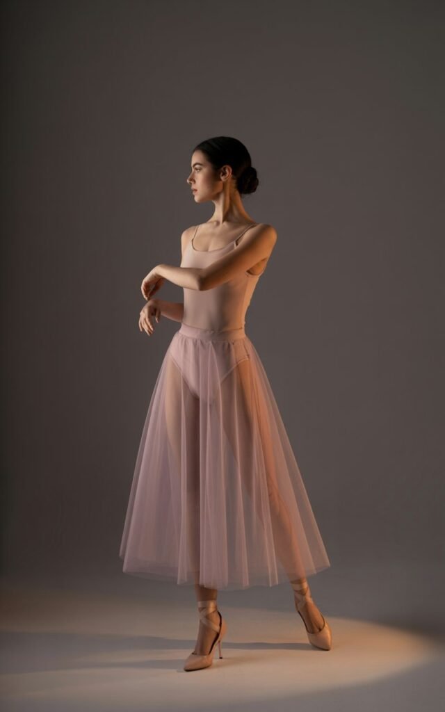 Studio setting with dramatic lighting. Model in a soft pink sheer tulle midi skirt layered over a nude fitted bodysuit, delicate heels. Hair sleek in a bun, elegant pose with arms lifted slightly, ballet-inspired.