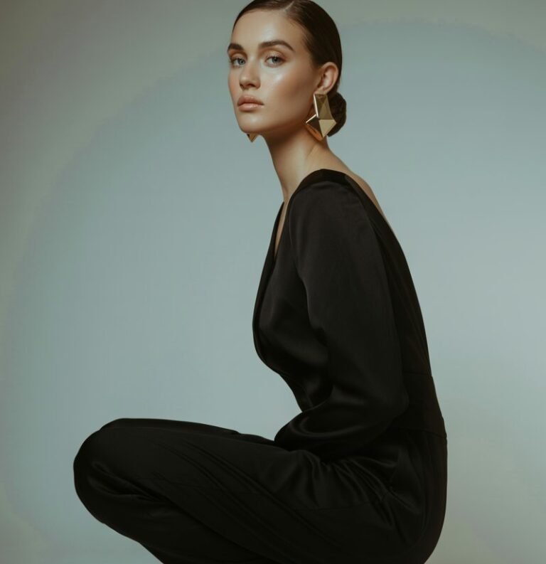 Studio setup with a soft gray backdrop. The model stands confidently, wearing a sleek black minimalist jumpsuit with bold gold earrings. She pairs it with strappy heels and a low bun. Lighting is soft and dramatic, focusing on her posture and accessories. The vibe is refined, clean, and editorial.