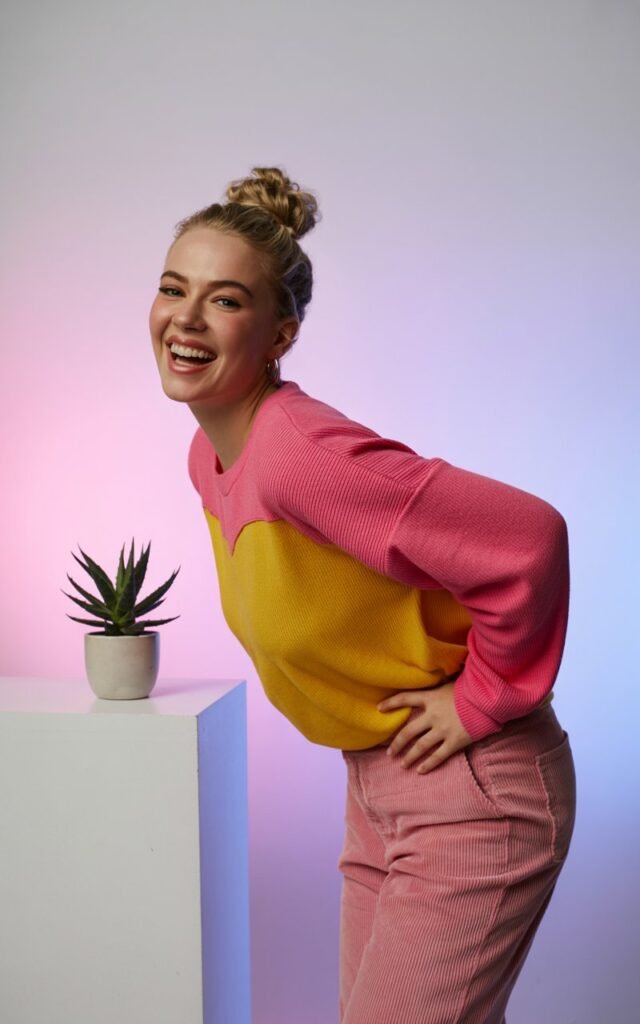 Studio shoot with pastel backdrops and soft lighting. She wears a bright color-block sweater with pink corduroy pants and white sneakers. Her hairstyle is a fun half-up bun, and she poses playfully with one hand on her hip, smiling big. Youthful and bold energy.