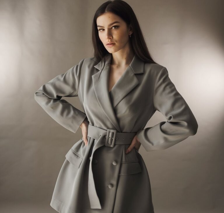 Studio shot of a white-skinned woman with long dark hair wearing a belted blazer dress and heels. Neutral taupe backdrop with soft spotlighting. She’s standing tall, one hand on her waist emphasizing the belt, gaze confident and empowering.