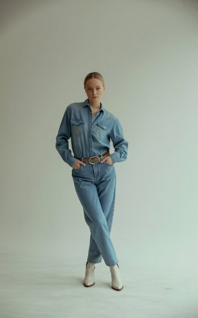 Studio shot with soft, diffused light highlighting a matching light-wash denim shirt and jeans set. The model accessorizes with a bold belt and white boots. She stands tall, hands in pockets, minimalist background emphasizing her sleek, modern cowgirl vibe.