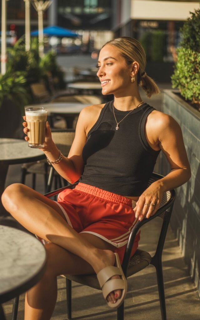 Urban café patio. Model in a black mock-neck tank and red track shorts, gold jewelry, and slides. Hair in a sleek ponytail, relaxed pose with iced latte. Soft golden-hour light highlighting her features.
