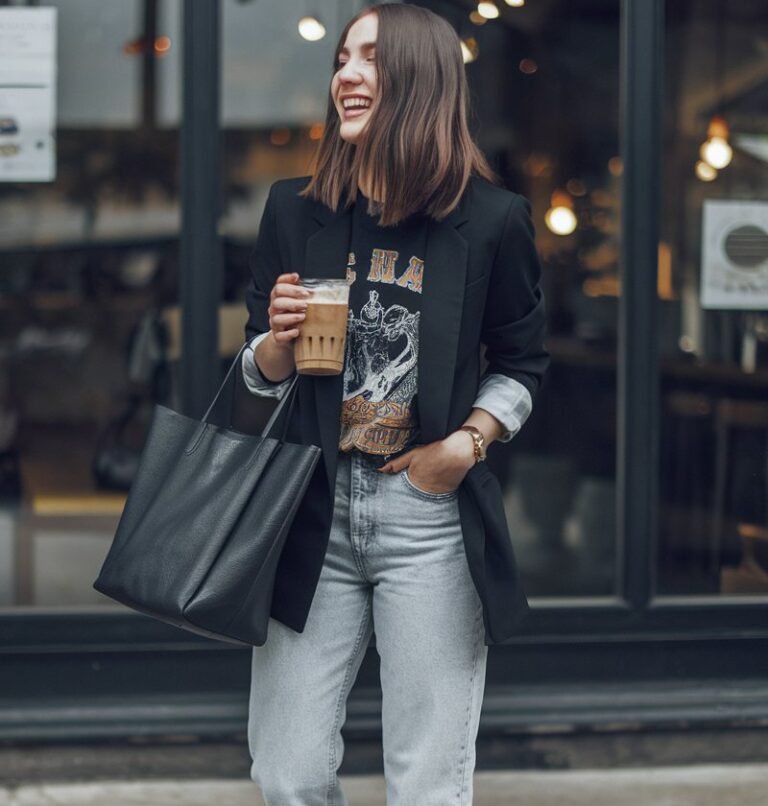 Urban coffee shop exterior scene. White-skinned brunette model with shoulder-length bob wears a black blazer, vintage band tee, and light-wash mom jeans. Paired with white sneakers and a tote bag. Natural daylight, candid mid-laugh pose while sipping iced coffee. Effortless cool-girl charm.