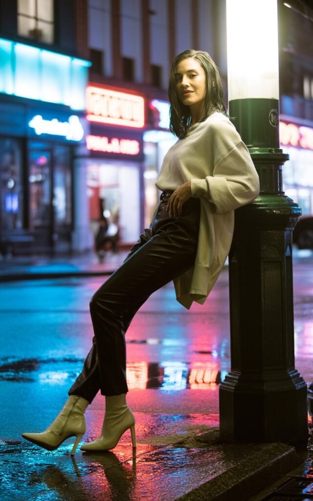 Urban night scene of a white-skinned woman with sleek black hair wearing fitted black leather pants, oversized cream sweater, and stiletto boots. Neon reflections on wet pavement create a cinematic mood. She’s leaning on a streetlight, half-smiling with a confident, bold pose.