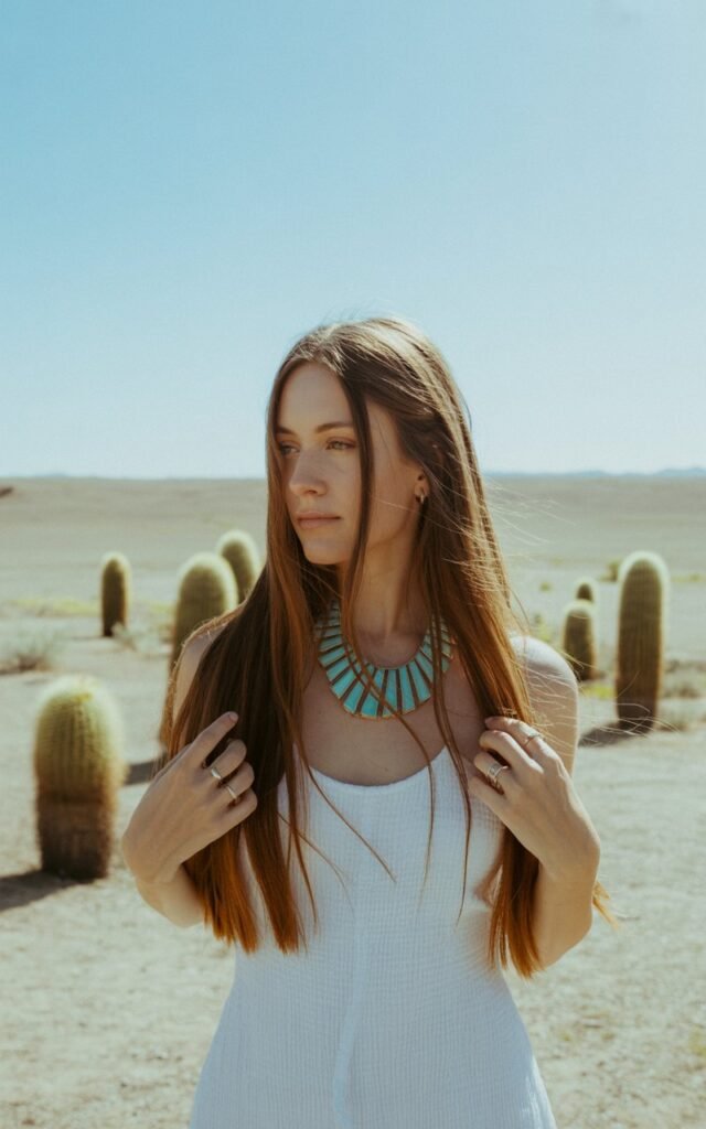White-skinned brunette woman with long straight hair, wearing a simple white cotton dress, turquoise statement necklace and rings. Shot in a sunlit desert backdrop with cactus plants. Natural daylight, clean composition. She’s standing gracefully with calm confidence, wind lifting her hair slightly.