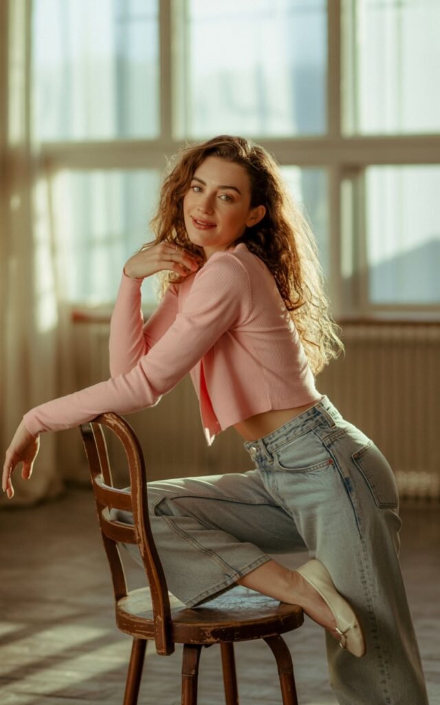 A cozy indoor studio with diffused window light. The model stands leaning slightly on a chair, wearing a cropped pastel cardigan with light-wash high-rise jeans and ballet flats. Her hair is loose and soft, expression gentle and approachable.