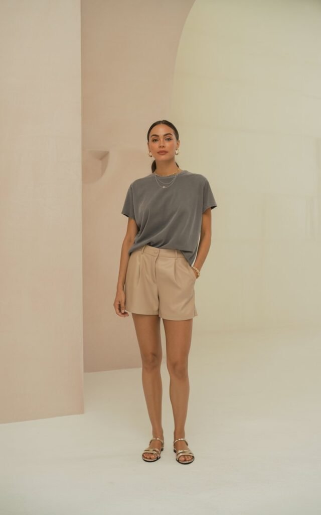 A minimalist studio with neutral tones. The woman stands with one hand in her pocket wearing a gray boxy tee tucked into beige tailored shorts, with simple sandals and delicate jewelry. Her hair is a low ponytail, makeup fresh and soft. The light is natural and clean.